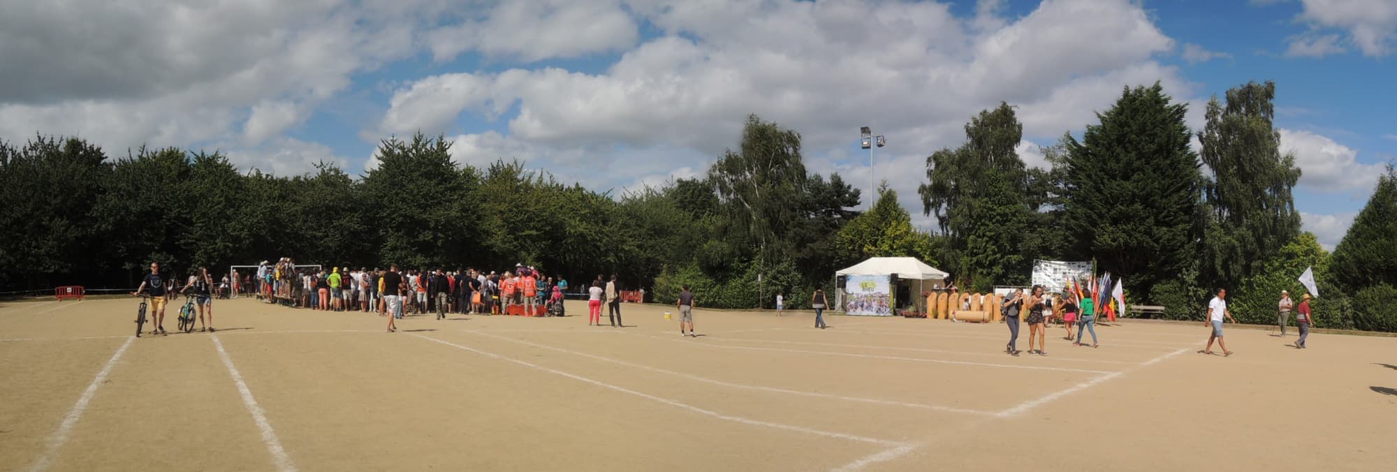Un large terrain de sable divisé en couloirs lors d'un tournoi de Mölkky, avec joueurs et spectateurs sur un côté.