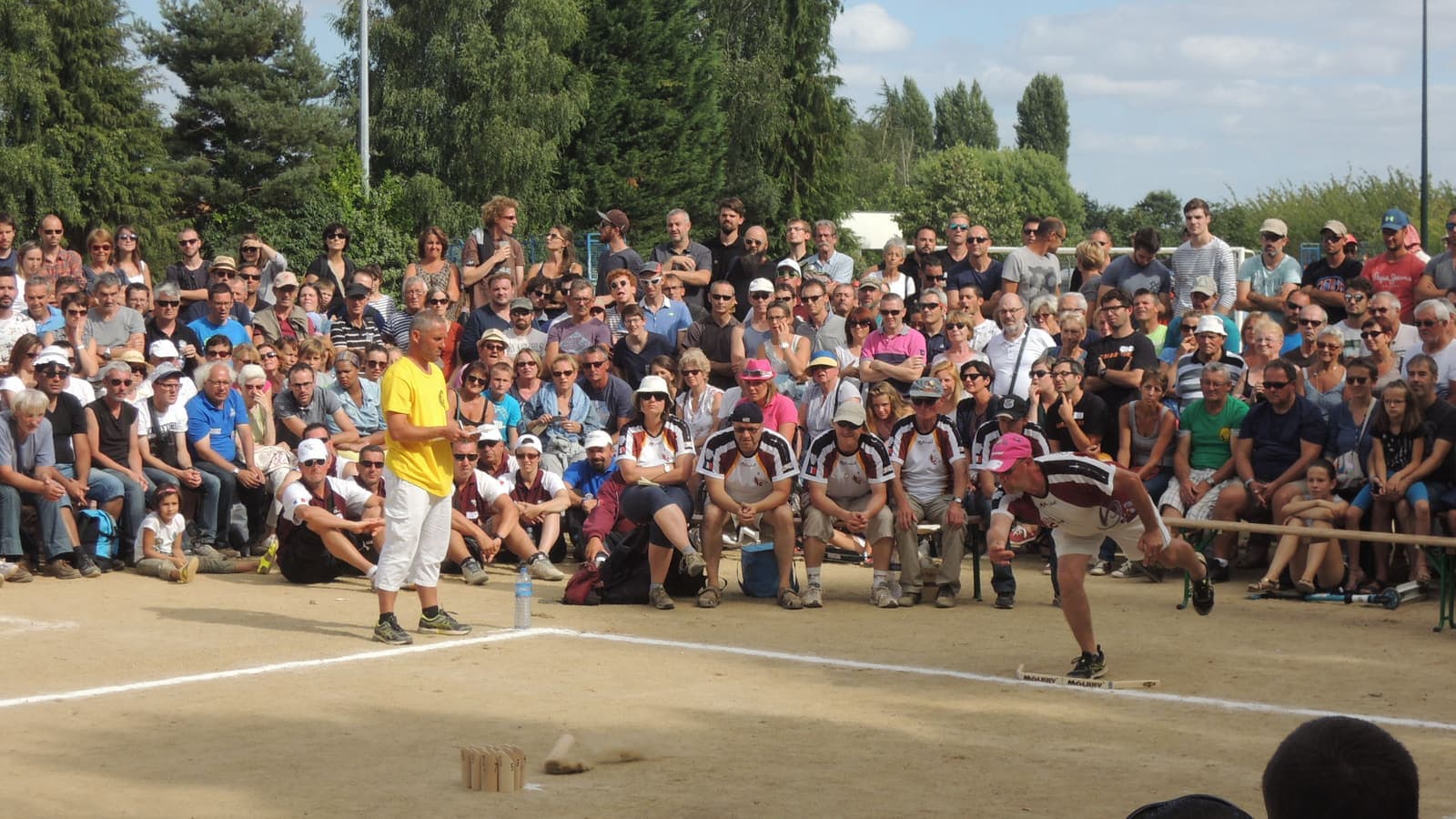 Un joueur vient de lâcher le mölkky par en dessous ; le bâton est en l'air au-dessus du sable, les douze quilles se dressent à l'autre bout et le public regarde.