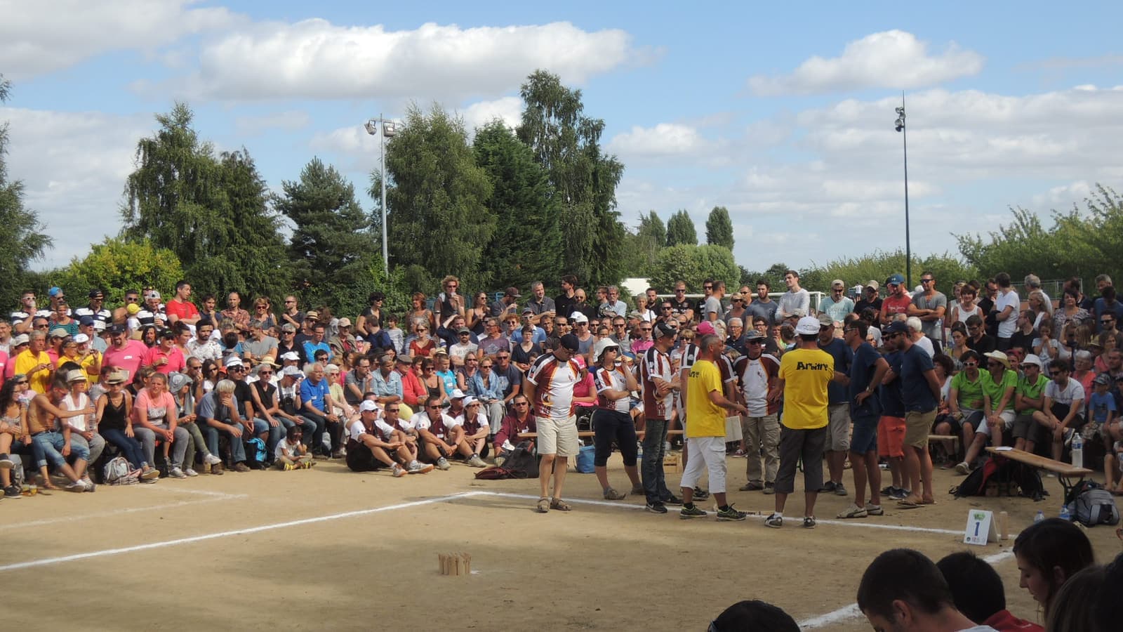 Une foule nombreuse, debout et assise autour d'un terrain de sable lors d'un tournoi de Mölkky, regarde deux équipes et un arbitre près de la ligne de lancer.